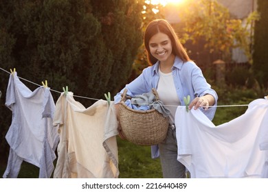 Smiling Woman Holding Basket And Hanging Clothes With Clothespins On Washing Line For Drying In Backyard