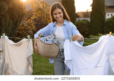 Smiling woman holding basket and hanging clothes with clothespins on washing line for drying in backyard - Shutterstock ID 2271602853