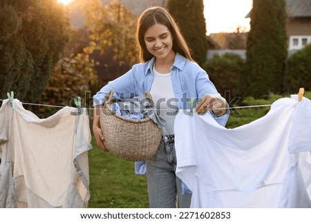 Smiling woman holding basket and hanging clothes with clothespins on washing line for drying in backyard