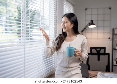 Smiling woman holding a blue cup while looking through window blinds in a modern home office with natural light and comfort. - Powered by Shutterstock