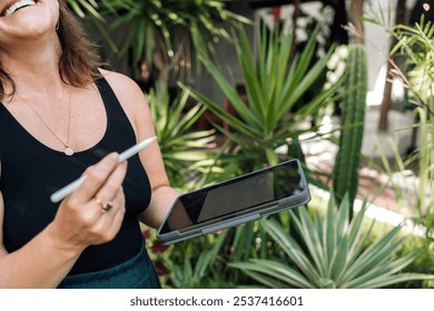 Smiling woman holding a digital tablet and stylus pen, standing outdoors surrounded by lush greenery. The relaxed, natural setting complements the joy of working creatively in nature - Powered by Shutterstock