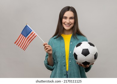 Smiling Woman Holding Flag Of United States Of America And Soccer Black And White Ball, United Soccer League, Wearing Casual Style Jacket. Indoor Studio Shot Isolated On Gray Background.