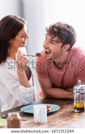 Smiling woman holding fork with waffle near boyfriend, tea and chocolate spread on blurred foreground