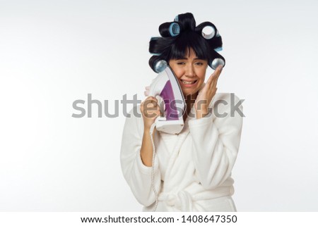 smiling woman holding hair curlers holding an iron on her face on a light background                               