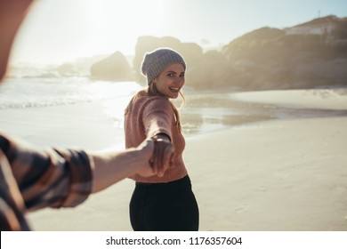Smiling woman holding man's hand while walking on the beach. Young couple walking along the beach. Happy young woman holding hand of boyfriend. Man following girlfriend walking. POV.