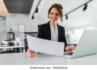Smiling woman holding one paper, while using laptop. - Shutterstock ID 1922042531