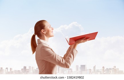 Smiling Woman Holding Open Notebook On Background Of Cityscape And Cloudy Blue Sky. Business Concept With Copy Space. Elegant Young Woman In White Business Suit Hand Up With Open Book.