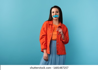 mujer sonriente sosteniendo un palo con bigote de papel sobre fondo azul