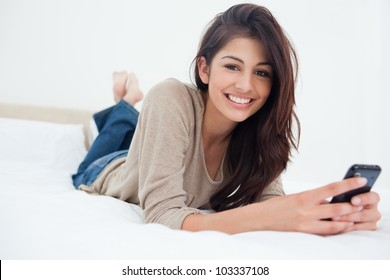 A smiling woman holds her phone in front of her as she looks forward while on the bed. - Powered by Shutterstock