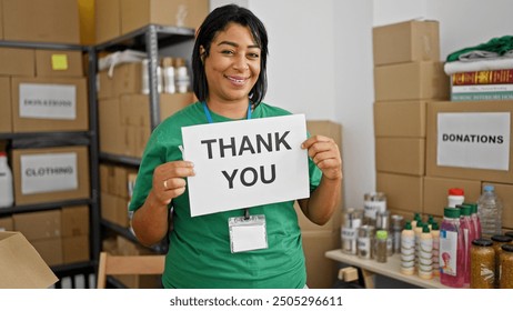 A smiling woman holds a 'thank you' sign in a donation center surrounded by boxes and supplies.