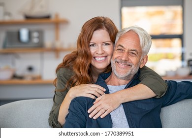 Smiling Woman Hugging Her Senior Husband On Couch From Behind In The Living Room. Loving Retired Couple Looking At Camera And Smiling At Home. Portrait Of Smiling Woman Embracing Mature Man On Sofa.