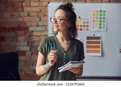 Smiling woman, interior designer or architect in casual wear with messy hairdo holding pen and notebook, looking aside, working on new creative project, standing near board with color samples - Shutterstock ID 2126792465