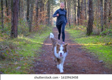 Smiling Woman Jogging Outdoors With Her Dog Running Ahead On A Dirt Trail In Forest Or Park