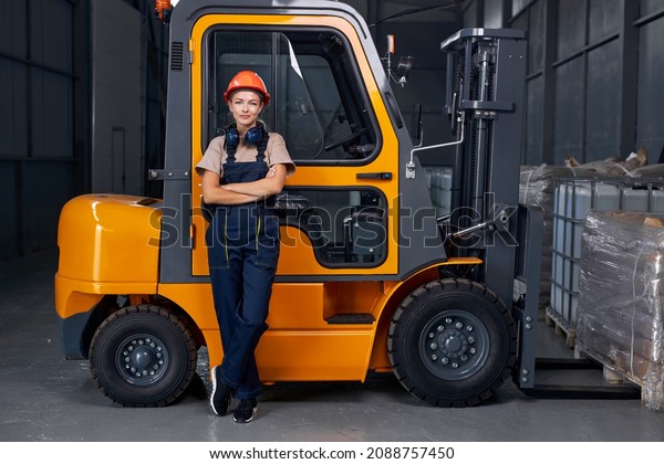 smiling woman labor worker near forklift driver in\
industry factory logistic shipping warehouse, posing with arms\
folded, dressed in uniform and orange protective hardhat, after\
successful good job
