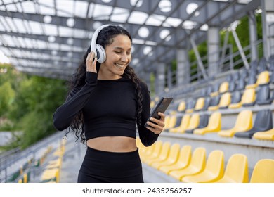 A smiling woman listens to music through white headphones, using her smartphone in an empty stadium seating area. - Powered by Shutterstock