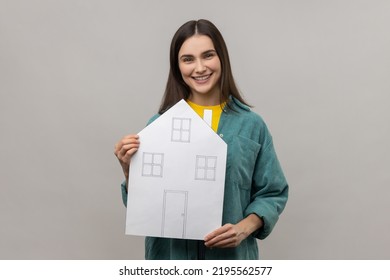 Smiling Woman Looking At Camera, Holding Paper House With Positive Expression, Dreaming Of Own Home, Wearing Casual Style Jacket. Indoor Studio Shot Isolated On Gray Background.