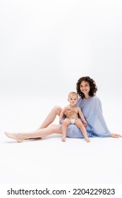 Smiling Woman Looking At Camera While Holding Baby Girl And Sitting On White Background