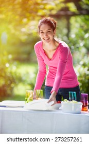 A Smiling Woman Is Looking At Camera While Setting The Table In The Garden For Her Family