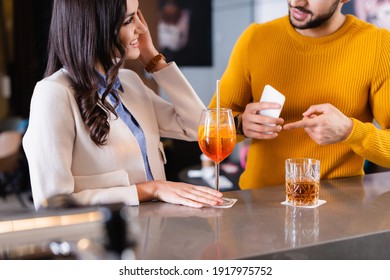 Smiling woman looking at friend pointing at smartphone near beverages on bar counter on blurred foreground