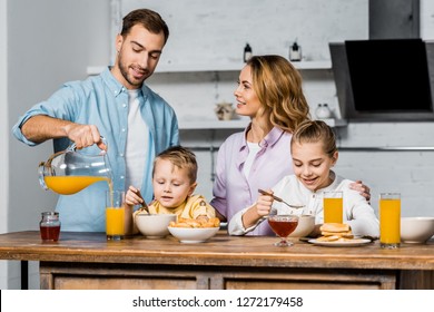 Smiling Woman Looking At Husband Pouring Orange Juice In Glass While Children Eating Oatmeal