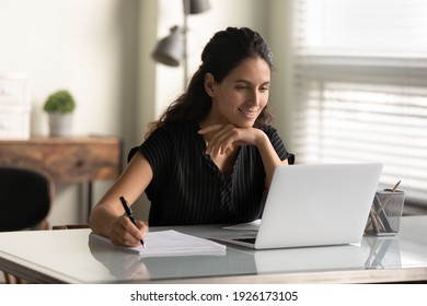 Smiling woman looking at laptop screen, watching webinar or lecture, online course, taking notes, sitting at desk, motivated young female student studying, businesswoman freelancer working on project