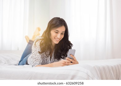 Smiling woman looking at mobile phone lying down on white bed. Happy brunette young woman using cellphone at home. Beautiful girl typing on smartphone and surfing the net.