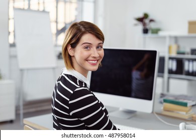 Smiling Woman Looking Over Shoulder While Working On Large Blank Computer Screen In Small Office