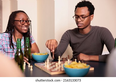 Smiling woman looking at pensive friend moving chess piece on board. Casual people relaxing by playing strategy boardgames while enjoying fun leisure time together at home in living room.