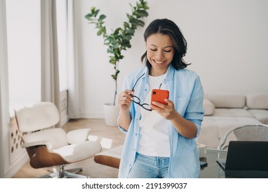 Smiling Woman Looking In Phone Screen In Office. Happy Female Businessperson Reading Pleasant Good News Message, Holding Smartphone And Glasses In Hands, Enjoying Chatting Communicating Online.