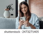 Smiling woman looking at smartphone screen, sitting on couch, enjoying leisure time with mobile device at home, happy young female chatting in social network or surfing internet, shopping online