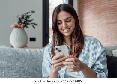 Smiling woman looking at smartphone screen, sitting on couch, enjoying leisure time with mobile device at home, happy young female chatting in social network or surfing internet, shopping online - Powered by Shutterstock