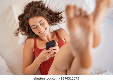 Smiling woman lying on white bed while lifting legs up and using app on smartphone. High angle view of beautiful young woman checking smart phone in morning. Top view of happy girl using mobile phone. - Powered by Shutterstock