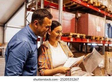 Smiling woman and man in casual clothes are working in warehouse. Young couple of employees laugh and mark presence of product storage in workshop. Workers check documentation and goods on shelves - Powered by Shutterstock