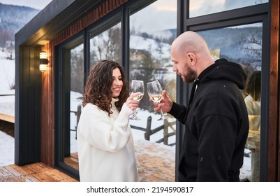 Smiling Woman And Man Clinking Glasses Of Wine And Celebrating Event Together On Winter Day. Couple In Love Toasting Enjoying Alcoholic Drink While Standing Outside Scandinavian House Barnhouse.