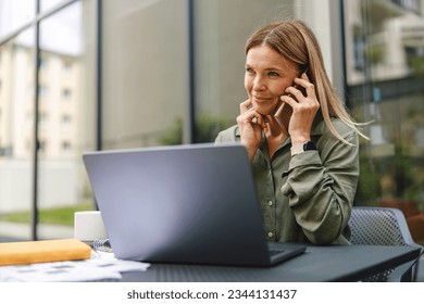 Smiling woman manager talking with client and work on laptop in cafe terrace. Distance work concept - Powered by Shutterstock