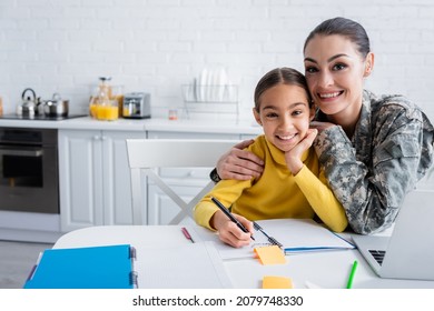 Smiling Woman In Military Uniform Hugging Child Near Notebooks And Laptop At Home