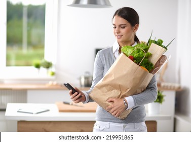 Smiling Woman With Mobile Phone Holding Shopping Bag In Kitchen