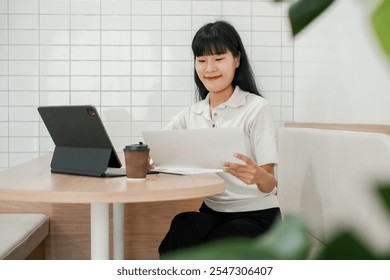 Smiling woman multitasking with tablet and documents in a stylish cafe, enjoying a coffee break. - Powered by Shutterstock