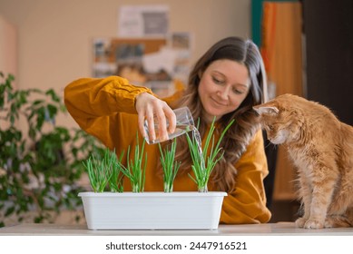 Smiling woman in a mustard yellow top waters green onion plants in a planter, while an inquisitive ginger cat watches closely. Growing greenery in the apartment, a mini vegetable garden. - Powered by Shutterstock