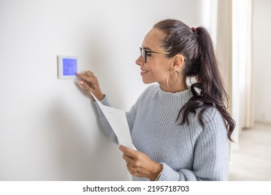 Smiling Woman Operates Digital Thermostat Installed On The Wall Inside Her Home.