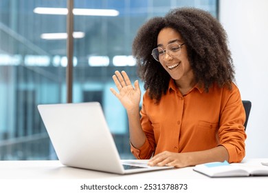 Smiling woman in orange shirt waves during online meeting, engaging in communication through laptop. Bright office setting conveys positive work-from-home environment and internet connectivity. - Powered by Shutterstock