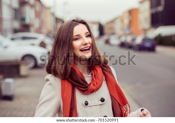 Smiling\
woman out at evening in the city looking away from camera, portrait\
in autumn spring wearing beige coat, red\
scarf