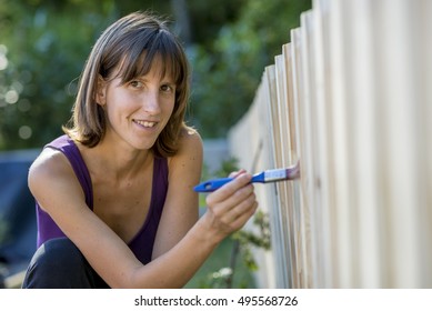 Smiling Woman Painting A Garden Fence With A Paintbrush In A Concept Of DIY And Yard Maintenance.