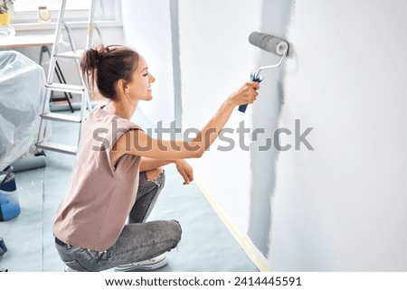 Smiling woman painting with paint roller on wall while crouching at home