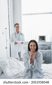 Smiling Woman In Patient Gown Near Blurred Doctor With Digital Tablet