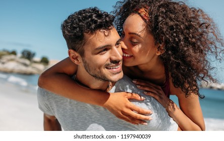 Smiling Woman Piggy Riding On Her Boyfriend And Kissing Him. Man Carrying His Girlfriend On His Back Outdoors.