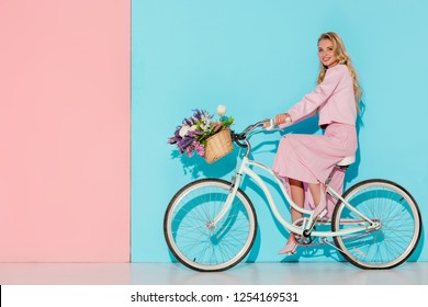 Smiling Woman In Pink Clothing Riding Bicycle With Flower Basket On Pink And Blue Background