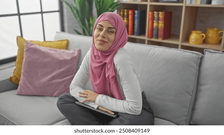 A smiling woman in a pink hijab lounging on a grey sofa indoors, exuding a comfortable and relaxed atmosphere - Powered by Shutterstock