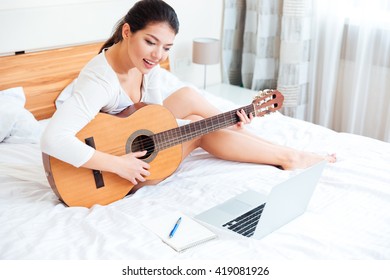 Smiling Woman Playing Records On Guitar Supported By Laptop Computer On The Bed At Home