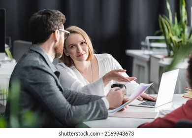 Smiling Woman Pointing With Finger Near Laptop And Colleague In Ad Agency